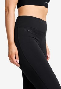 Kvinna i svarta högmidjade leggings och matchande crop top med RVRC-logotyp, stående mot en enkel vit bakgrund.