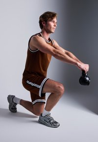 Barna sportos tank top és rövidnadrág fekete és fehér csíkokkal; egy fekete kettelbellt tart; szürke sportcipőt és fehér bokaszkókot visel.