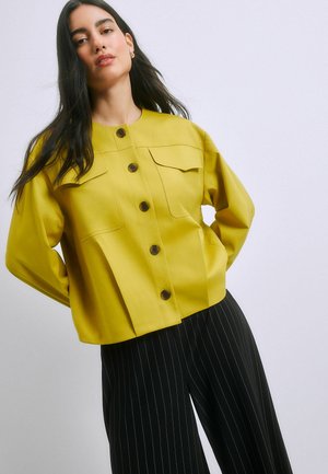 Woman with long dark hair wearing a mustard yellow buttoned jacket and black pinstriped pants against a plain light background.