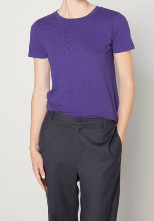 Osoba ubrana w dopasowaną, fioletową bluzkę z krótkim rękawem i ciemnoszare, dopasowane spodnie, jedna ręka w kieszeni, neutralne tło.
