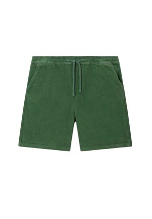 Homme portant un short en velours côtelé vert, des chaussettes blanches à rayures rouges avec le texte « OUT OF OFFICE », des chaussures bateau couleur camel et un t-shirt beige.