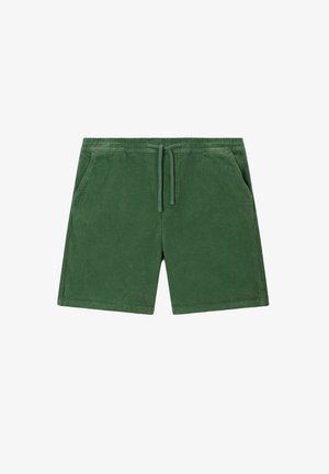 Homme portant un short en velours côtelé vert, des chaussettes blanches à rayures rouges avec le texte « OUT OF OFFICE », des chaussures bateau couleur camel et un t-shirt beige.