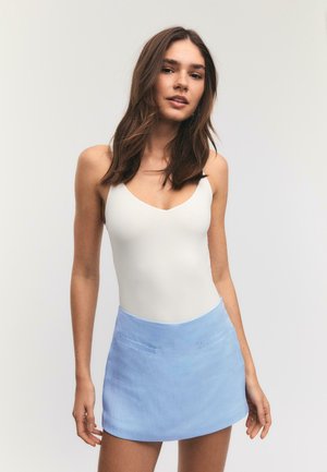 Femme aux cheveux bruns portant un haut blanc sans manches et une mini-jupe bleu clair, debout devant un fond gris uni.