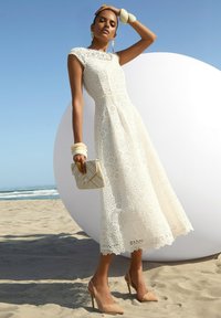 Weißes Spitzen-Midikleid mit floralem Muster, kurzen Ärmeln und tailliertem Oberteil. Kombiniert mit beigen Stiletto-Heels und einer dekorativen Clutch.