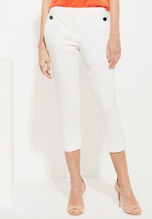 Woman wearing white cropped pants with black button details and nude high-heeled sandals, paired with an orange top.