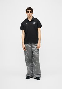 Νεαρός άνδρας που φοράει μαύρο πόλο BMW Motorsport, γκρίζο ανακλαστικό παντελόνι, μαύρα παπούτσια και στρογγυλά σκούρα γυαλιά ηλίου, στέκεται μπροστά σε λευκό φόντο.