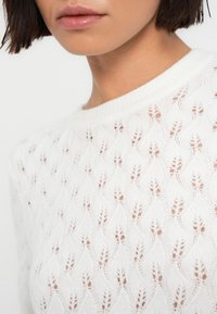 Λευκό πλεκτό πουλόβερ με υφή μοτίβου, που περιλαμβάνει σχέδια σε μορφή κυμάτων και μικρές ανοίγματα. Ο λαιμός είναι στρογγυλός, και το υλικό φαίνεται μαλακό.