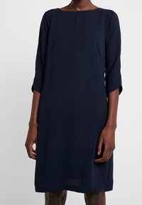 Navyblaues Kleid mit geradem Schnitt, rundem Ausschnitt, Dreiviertelärmeln und einem kleinen Schlitzdetail auf der Vorderseite. Glatte Stofftextur.