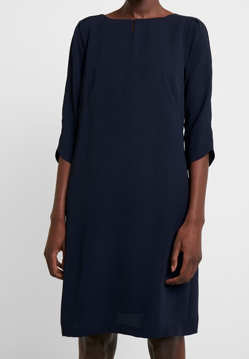 Navyblaues Kleid mit geradem Schnitt, rundem Ausschnitt, Dreiviertelärmeln und einem kleinen Schlitzdetail auf der Vorderseite. Glatte Stofftextur.