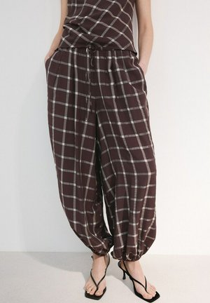 Femme portant un pantalon large marron à motif quadrillé blanc, avec des poignets élastiques et des sandales noires à talons hauts, les mains dans les poches.