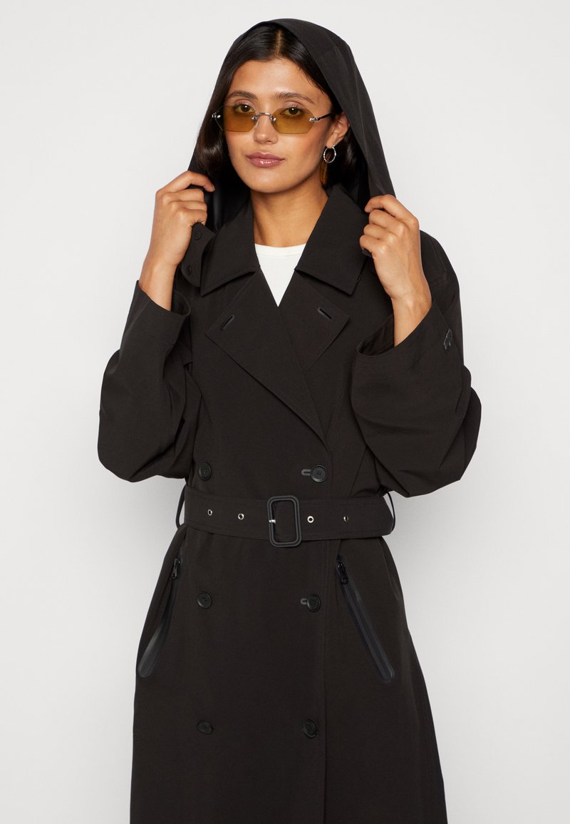 Lungo cappotto nero con design doppiopetto, ampio colletto, cintura in vita e cappuccio. Il modello indossa occhiali colorati e tiene il cappuccio.