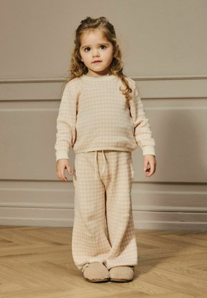 Jong kind met krullend haar, gekleed in beige geruite loungekleding en pluizige slippers, staande op een houten vloer tegen een beige muur.