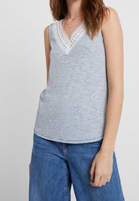 Koszulka bez rękawów w niebiesko-białe paski, z dekoltem w kształcie litery V wykończonym koronką i luźnym krojem, zestawiona z jasnoniebieskimi dżinsami.