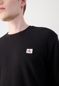 Young person wearing a black ribbed Calvin Klein sweater with a small red and white logo patch on the chest.