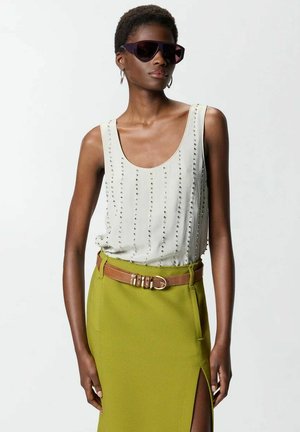 Donna che indossa un top bianco senza maniche con borchie, gonna verde oliva con spacco laterale, cintura marrone, grandi occhiali da sole scuri e orecchini a cerchio.