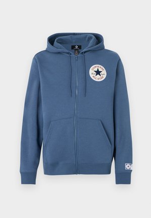 Kék cipzáros pulóver puha anyagból, bal mellrészen kerek logófolttal és első zsebekkel. Kapucni húzózsinórokkal.