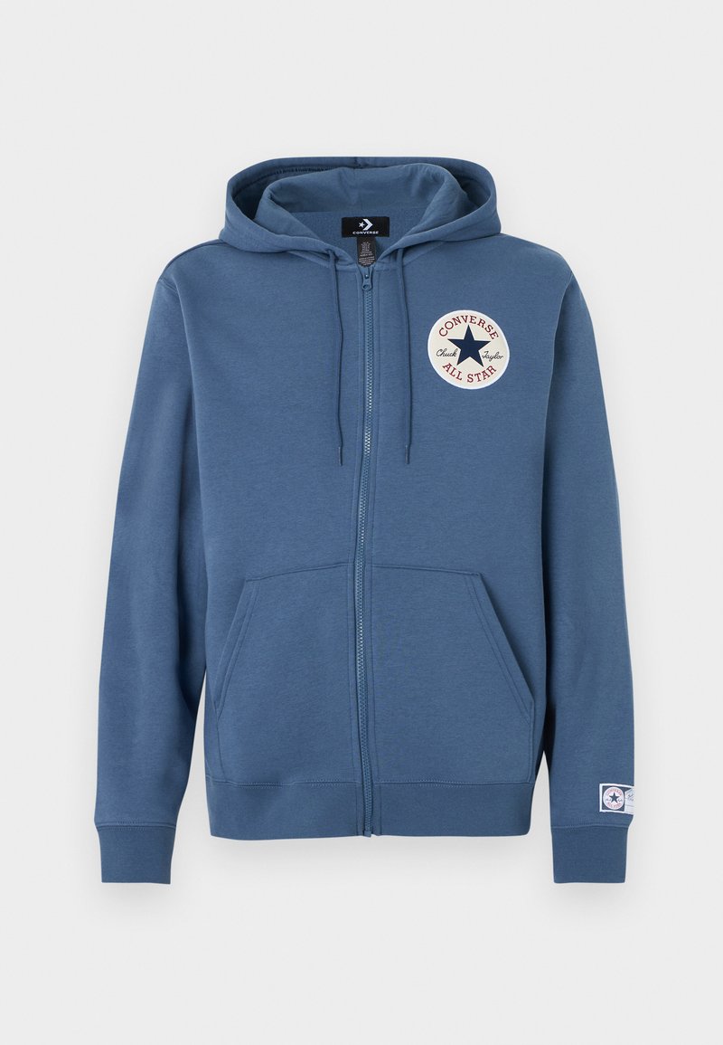 Blå lynlåshoodie lavet af blødt stof, med et rundt logopatch på venstre bryst og forlommer. Hætte med snore.