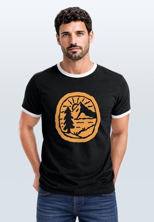 Mann mit kurzen lockigen Haaren und Bart, trägt ein schwarzes T-Shirt mit weißem Rand und orangefarbenem Berg- und Baum-Motiv, Hände in den Hosentaschen der Jeans.
