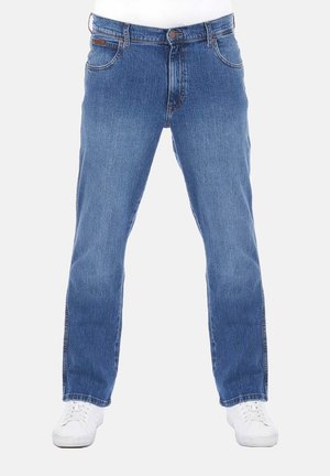 Gerade geschnittene blaue Jeans aus Denim, getragen mit weißen Turnschuhen, Frontansicht vom Taille bis zu den Füßen vor einfachem Hintergrund.