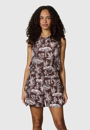 Kvinde med krøllet hår iført brun ærmeløs romper med hvide tropiske træ- og plante mønstre, hænderne i lommerne, stående mod grå baggrund.
