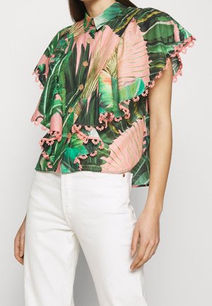 Ärmellose Knopfleiste-Bluse mit mehrlagigen Rüschen, tropischem grün-rosa Blättermuster und pinkem Pomponbesatz, getragen mit weißen Hosen.