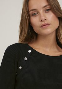 Schwarzer gerippter Pullover mit weiter Ausschnittöffnung und silbernen Knopfdetails an der linken Schulter, die einen texturierten Kontrast schaffen.
