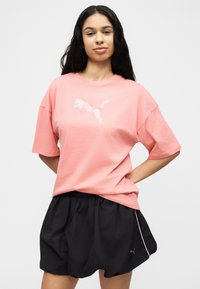 Jeune femme aux longs cheveux bruns portant un t-shirt rose ample de la marque Puma et un short noir, debout les mains sur les hanches devant un fond uni.