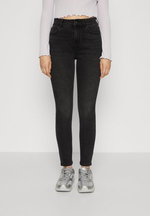 Vrouw draagt zwarte skinny jeans met hoge taille, zilveren sneakers en een wit cropped top met lange mouwen en gegolfde randen.