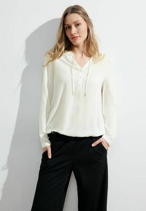 Chemise blanche à manches longues avec une capuche, dotée d'une patte de boutonnage et de finitions en cordon vert, portée avec un pantalon noir à taille élastique.