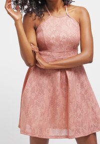 Rosa Spitzenkleid mit Halterneck, tailliertem Oberteil und weit ausgestelltem Rock. Mit floralen Mustern, dünnen Trägern und einem transparenten Überwurf.