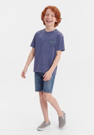 Rudowłosy chłopiec uśmiechający się i stojący, ubrany w wyblakły fioletowy T-shirt z logo GAP, dżinsowe szorty oraz szare buty wsuwane.
