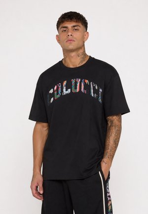 Man met tattoos op nek en arm draagt een zwart T-shirt met kleurrijk patroon en de tekst "COLUCCI", staand tegen een effen achtergrond.