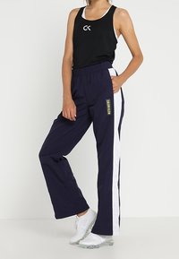 Frau in schwarzem ärmellosem Oberteil und dunkelblauen Trainingshosen mit weißen Streifen sowie weißen Turnschuhen, stehend vor einem weißen Hintergrund.