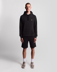 Čierna mikina s kapucňou a klokaním vreckom, so žltým logom, skombinovaná s čiernymi šortkami. Sivé a béžové tenisky, biele ponožky, neutrálne pozadie.