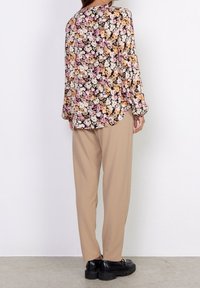 Blusa floreale a maniche lunghe con colori vari su uno sfondo scuro, abbinata a pantaloni beige chiaro e scarpe nere spesse.