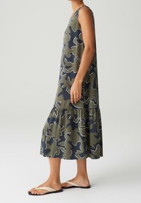 Femme debout de profil portant une robe midi sans manches, ample, avec un motif abstrait vert et bleu marine et des sandales beiges.