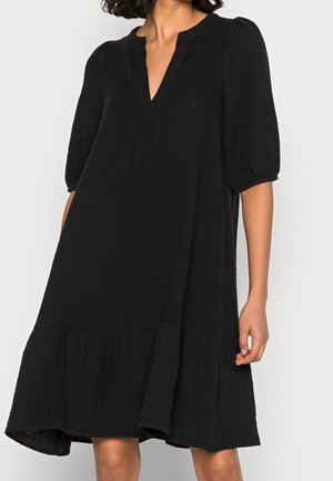 Femme portant une robe noire ample mi-longue aux manches bouffantes jusqu'au coude et un décolleté en V avec des plis subtils.