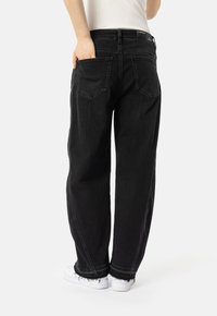 Isik, kes kannab lahtisi musti denim-pükse, millel on toored servad, ja valgeid tennisetossusid, käsi tagataskus, seismas valgel taustal.