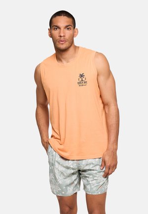 Hombre con camisa sin mangas color melocotón con logo de palmera y pantalones cortos de estampado floral verde claro, de pie con una mano en el bolsillo.