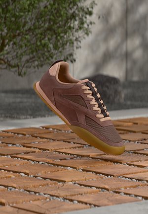 Bruine suède en mesh sneaker met beige veters die boven een houten blok en kiezelige buitenoppervlakte zweeft.