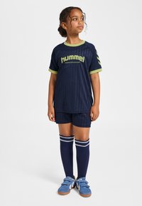 Barnens marinblå sportoutfit med nålrandiga ränder, gröna accenter och en "hummel"-logotyp. Inkluderar shorts, knähöga strumpor och blå skor.