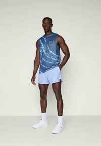 Modrá tie-dye bezrukávová blúzka s bielym textom v kombinácii s svetlomodrými šortkami. Model má na sebe biele tenisky a ponožky, stojí na jednofarebnom pozadí.