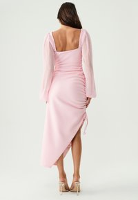 Woman wearing a pink asymmetrical dress with puffed long sleeves and ruched side, standing barefoot against a plain light background.