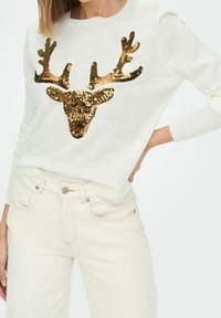 Crèmekleurige trui met een design van een pailletten hertenkop, met gouden takken aan de voorkant, gedragen met lichtgekleurde jeans. Zachte textuur.