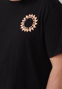 Zwarte t-shirt met een ronde zonnebloemontwerp in perzik, gesitueerd op de linkerkant van de borst. Gemaakt van zachte, gestructureerde stof.