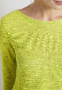 Pull en tricot vert citron avec un large décolleté, présentant un motif rayé texturé. Le tissu est léger et semi-transparent.
