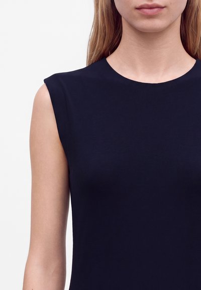 Gros plan d'une femme portant un haut noir sans manches avec un col rond sur un fond blanc uni.