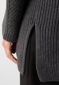 Gros plan d’une main à côté d’un pull en maille côtelée épaisse gris foncé avec une fente latérale, porté sur un tissu noir texturé.