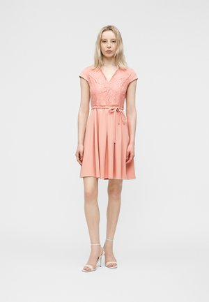 Kvinde står iført en kortærmet, knælængde lyserød kjole med blondeoverdel og bindebånd i taljen, kombineret med hvide sandaler med høje hæle.