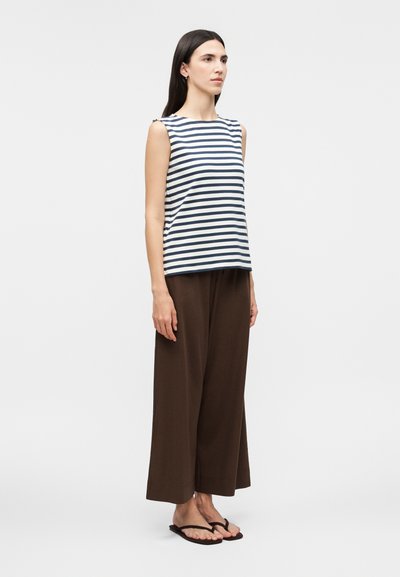 Femme aux longs cheveux foncés portant un haut sans manches rayé bleu marine et blanc, un pantalon large marron et des tongs noires, debout sur un fond blanc.